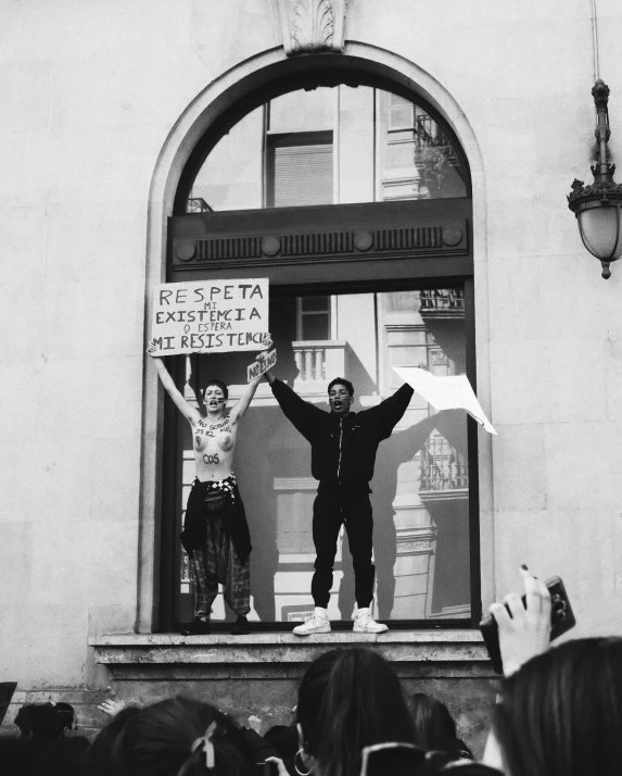 two people at a protest waving from a window