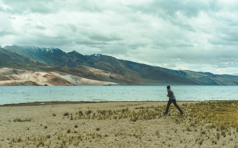 man walking through grass near a mountain lake