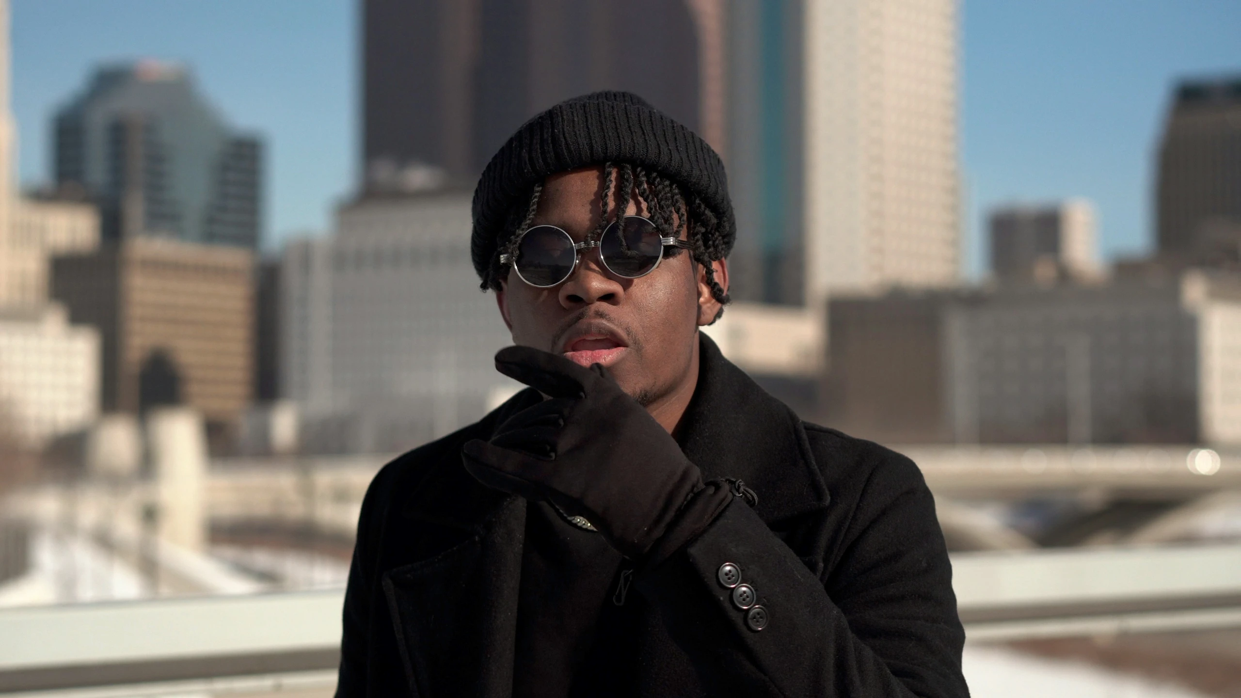 a young man with sunglasses, cap and scarf standing on a bridge with a cityscape in the background