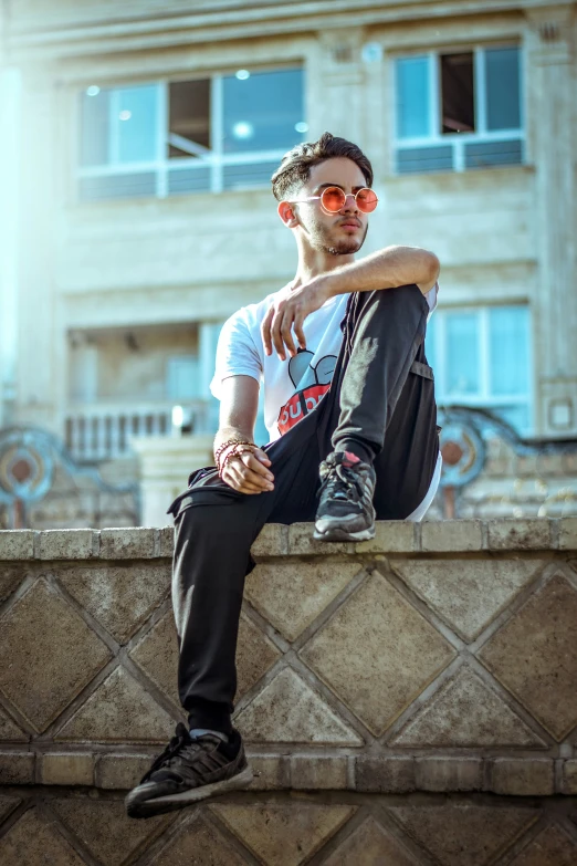 a young man wearing sunglasses and a t - shirt sits on a wall with his leg crossed