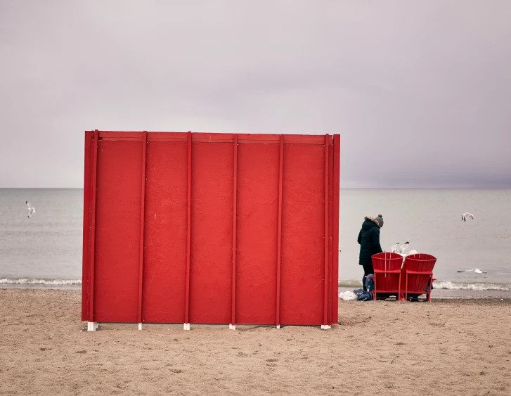 a tall red sculpture on the beach with people standing around it