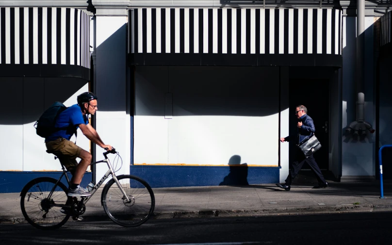 a man rides a bicycle past a building as a pedestrian walks by