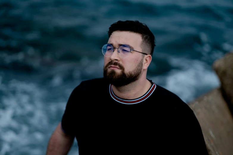 a man with glasses on standing next to a wave in the ocean