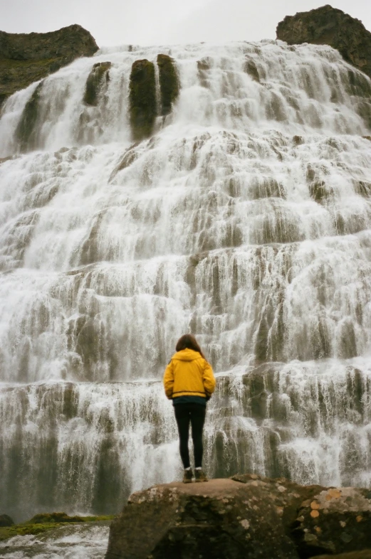 a woman in yellow jacket standing on rock in front of waterfall
