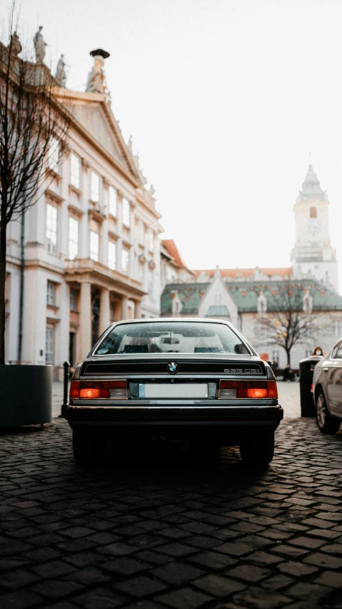 a black bmw e34 sits in the middle of a cobblestone road with other parked cars