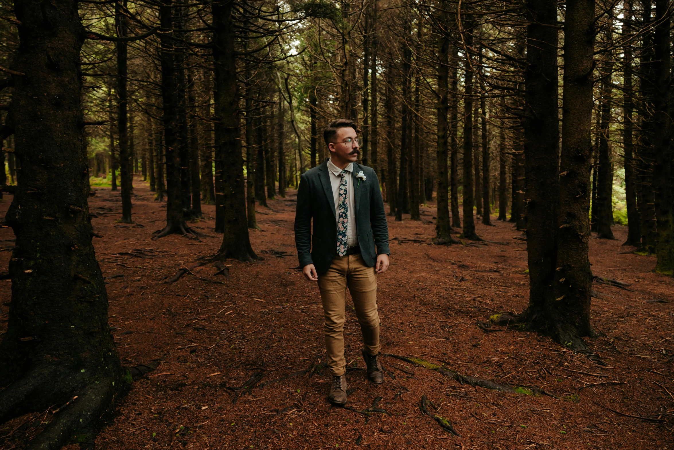 a man in a suit stands next to trees in the woods