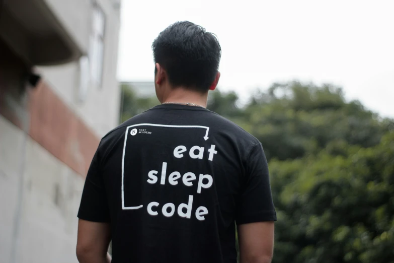 a man stands near a building wearing a black eat sleep code shirt