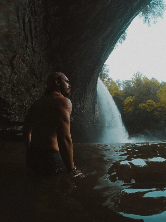 a shirtless man in front of a waterfall looking out at water