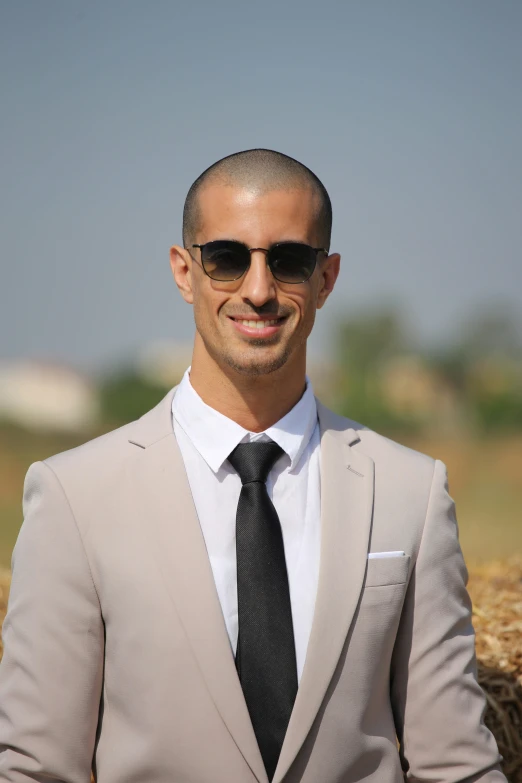 a well dressed man in a tan suit and sunglasses smiles at the camera