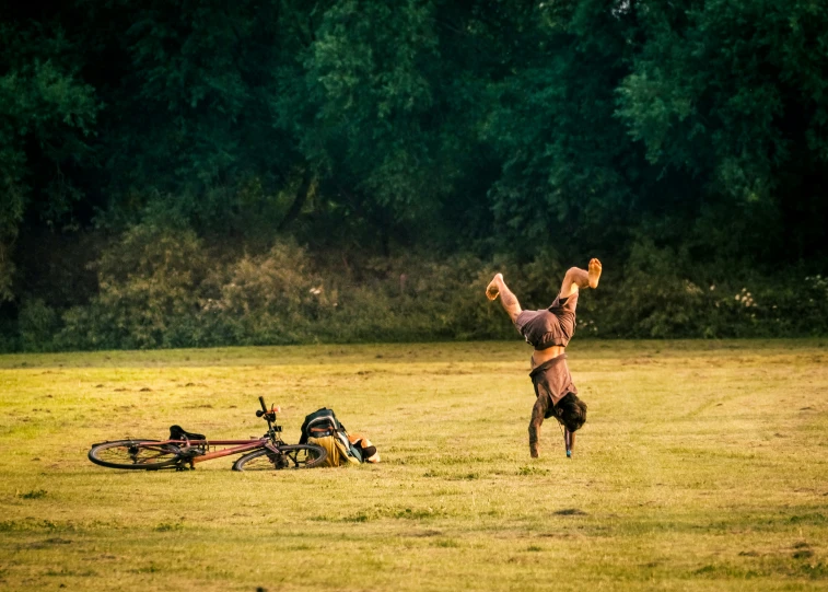 man with a hat doing handstand in a field next to bicycle