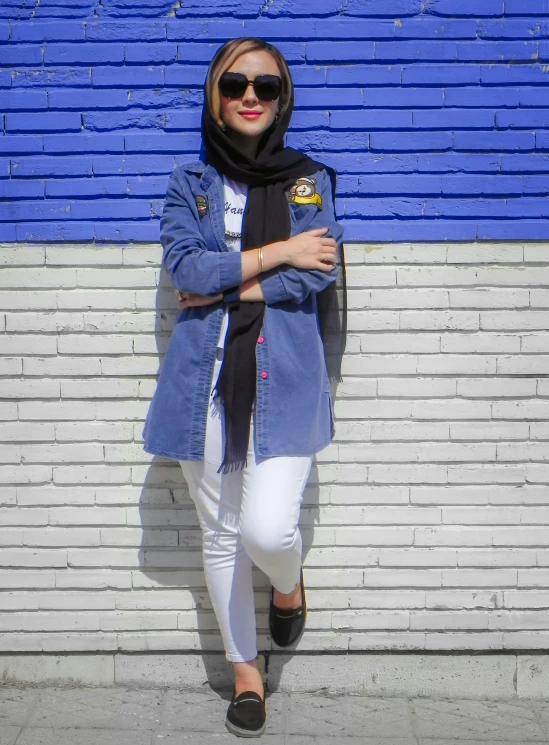 woman wearing white jeans and a denim jacket, standing against a brick wall with her hands on her hip