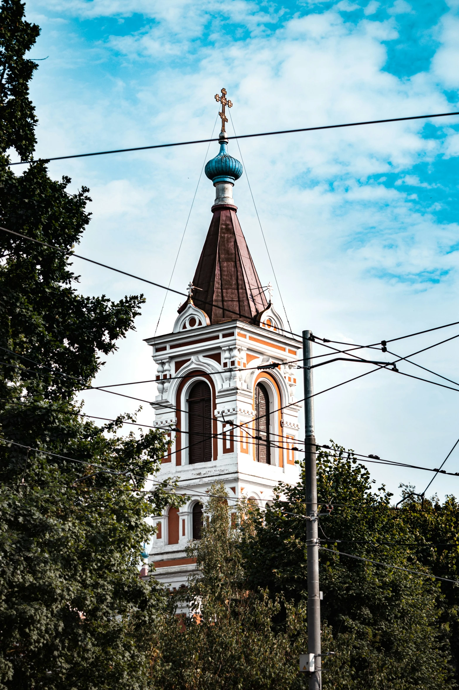 a white tower with a cross on it under blue skies and a tree in front of power lines