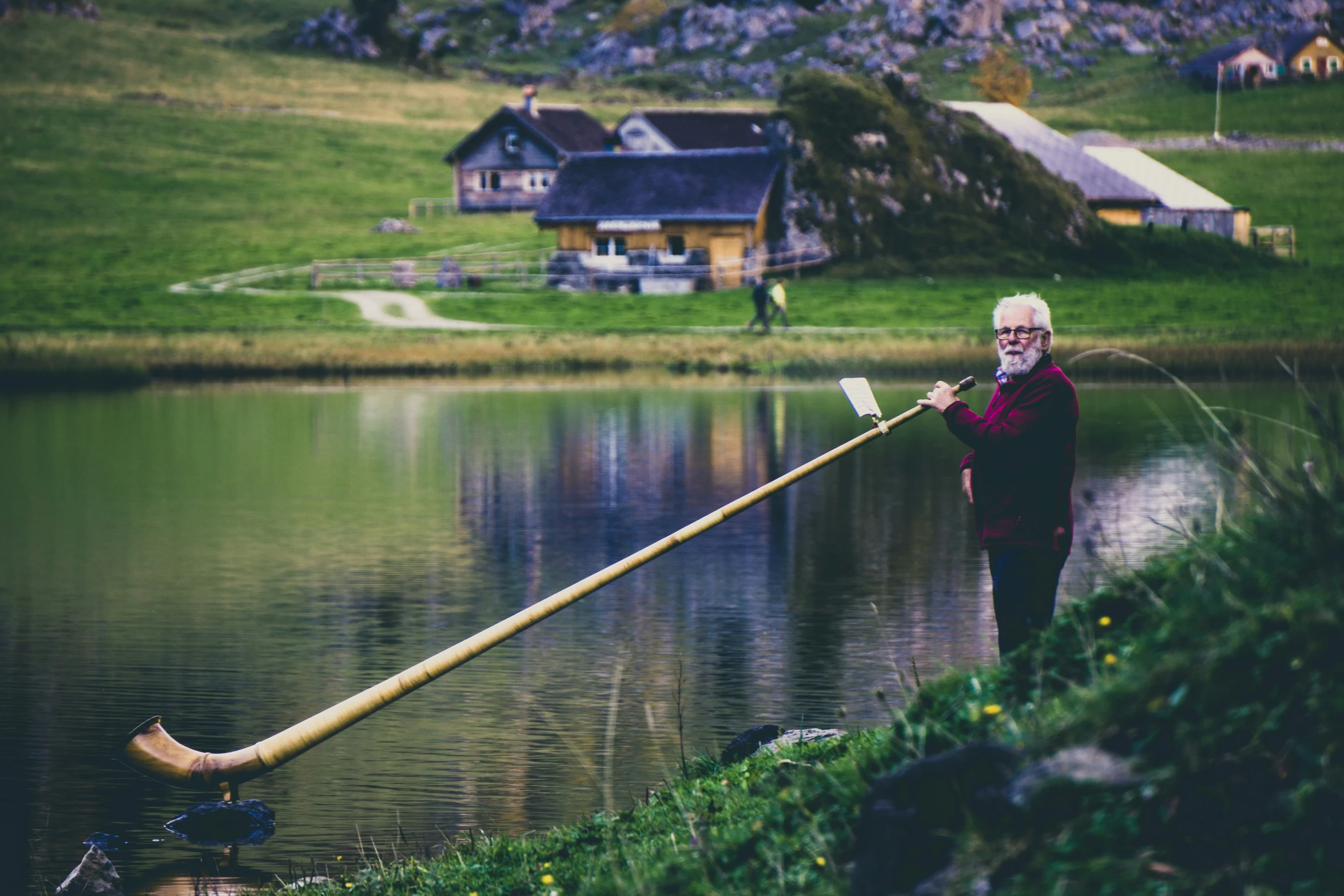 an elderly man holds a pole over water while a house sits in the background