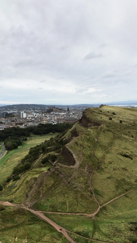 a view of a green hill with dirt winding it's sides and a large city in the distance