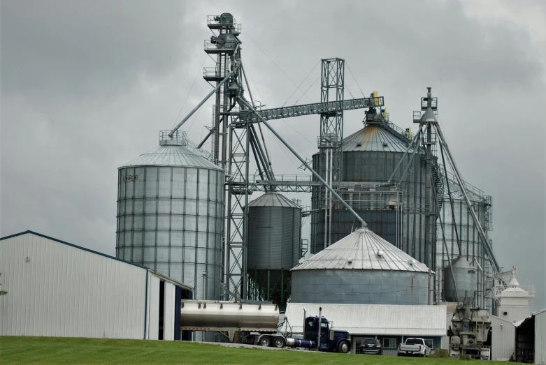 several gray silos, and one white truck are in a yard with a grey sky