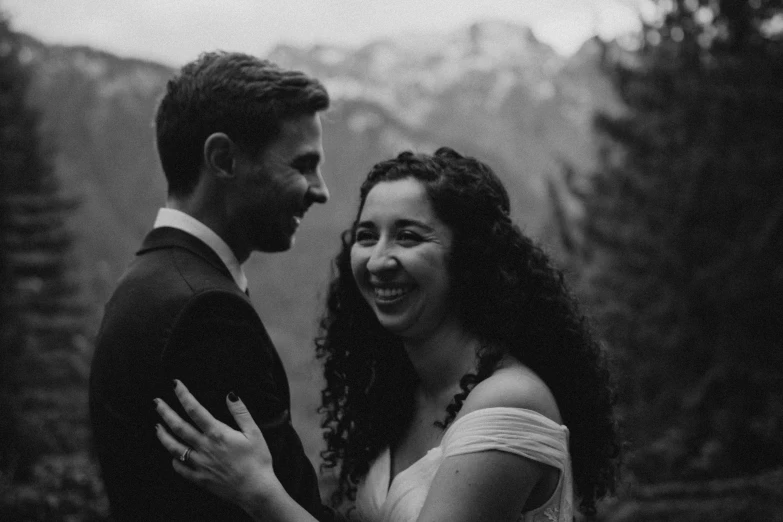 a smiling young couple stand on top of a hill looking into each others eyes