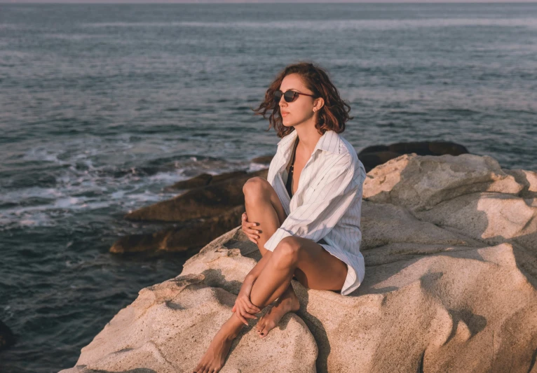 a woman with sunglasses on sitting on rocks by the ocean