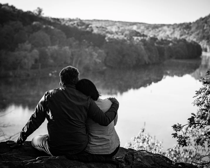 a couple sitting on the ground facing out at a lake