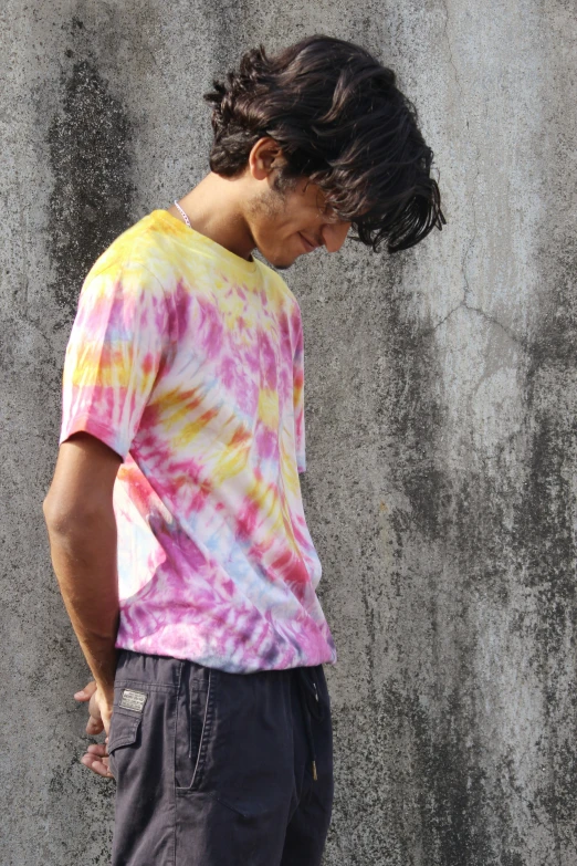 a man wearing a tie dye t - shirt stands against a concrete wall