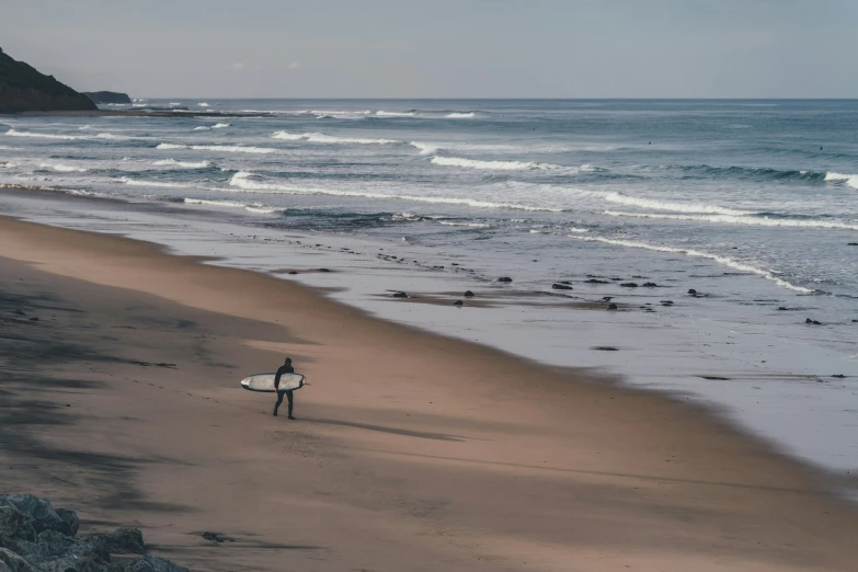 a lone surfer walks along a beach with waves crashing and rocky shore