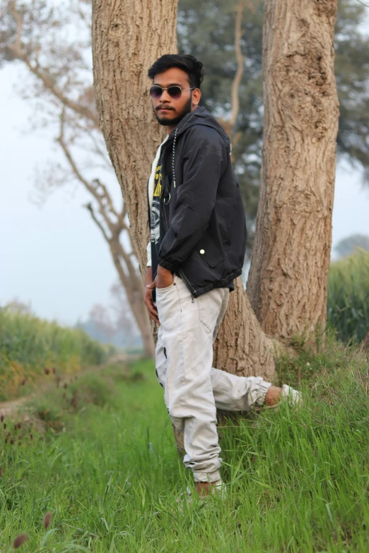 an indian man leaning against two trees wearing shades