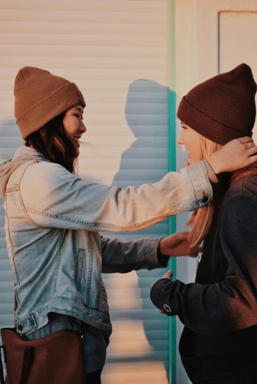 two people, one wearing a hat and the other wearing a denim jacket, one standing and holding another