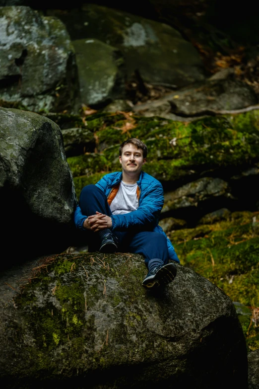 a man in a blue jacket and jeans sits on rocks