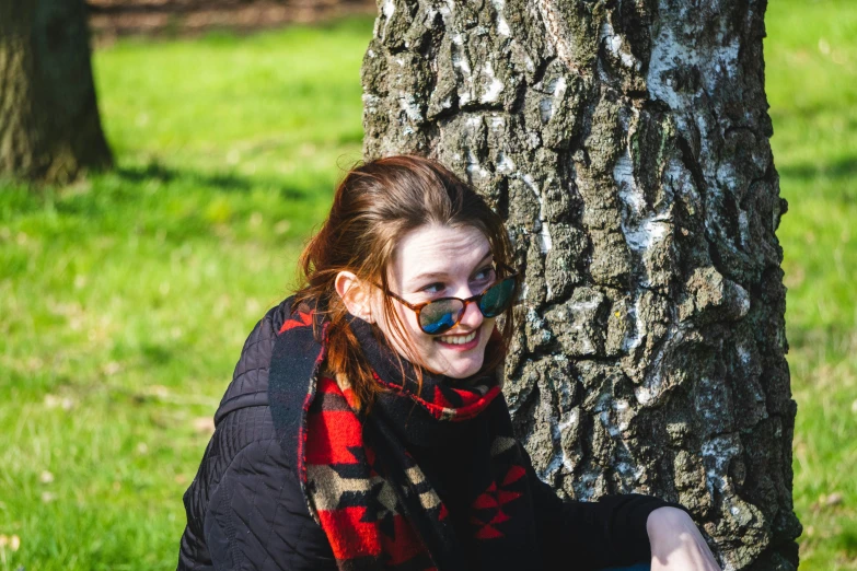 a girl with glasses sitting in the grass next to a tree