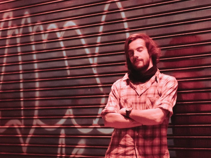 bearded man with arms crossed leaning against a wall with graffiti on it