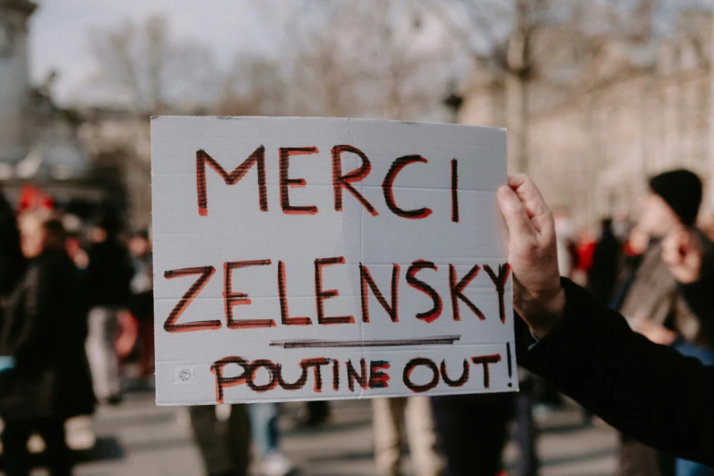 person holding up a sign saying merci zelensky while others gather on the street