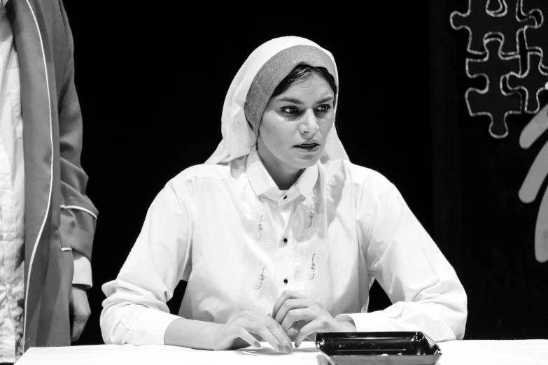 woman sitting at desk in white dress on stage with electronic device