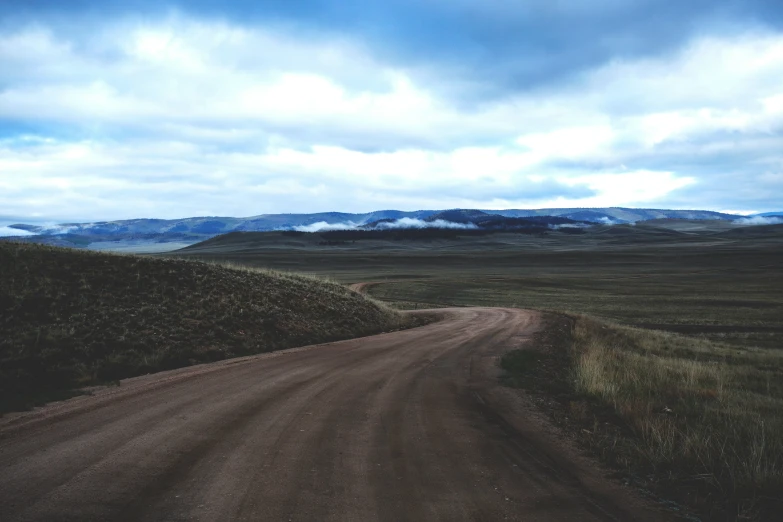 a dirt road is lined with thick grassy hills and rolling clouds