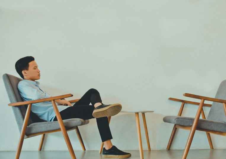 a man in black pants is sitting on a chair and a person is leaning against a wall with his feet up