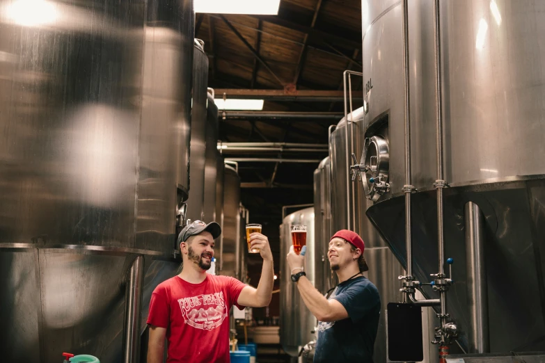 two men are standing in a brewery and one is holding a beer