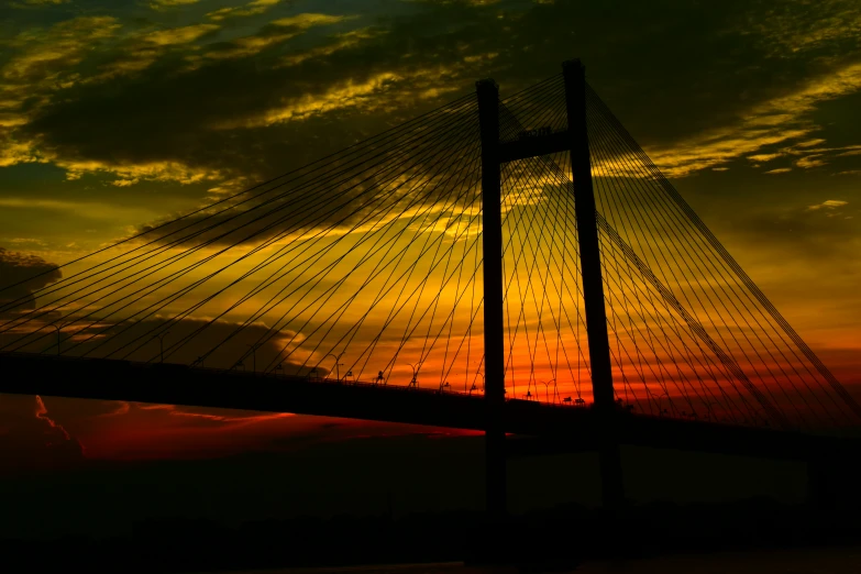 the sun sets on a cloudy day behind a suspension bridge