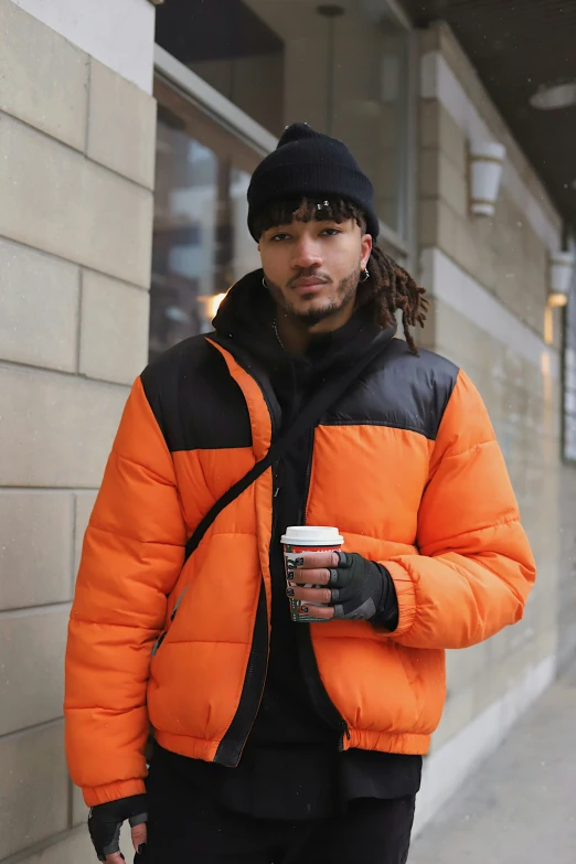 a man in an orange and black puffer jacket stands next to a building while holding a cup