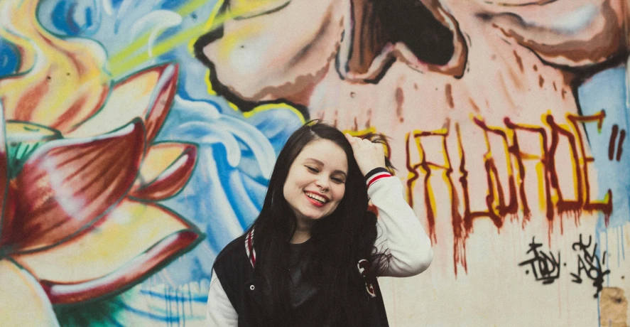 a smiling woman leans against a wall covered with graffiti