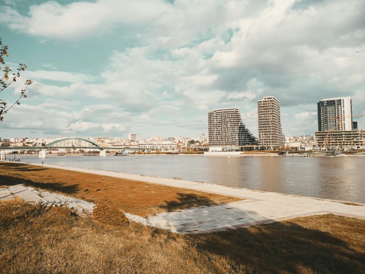 the view from a waterfront park shows tall buildings, an old bridge and a cloudy sky