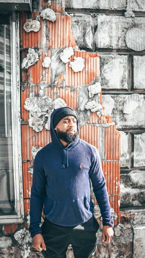 a man with a hoody and a beard poses in front of a dilapidated window