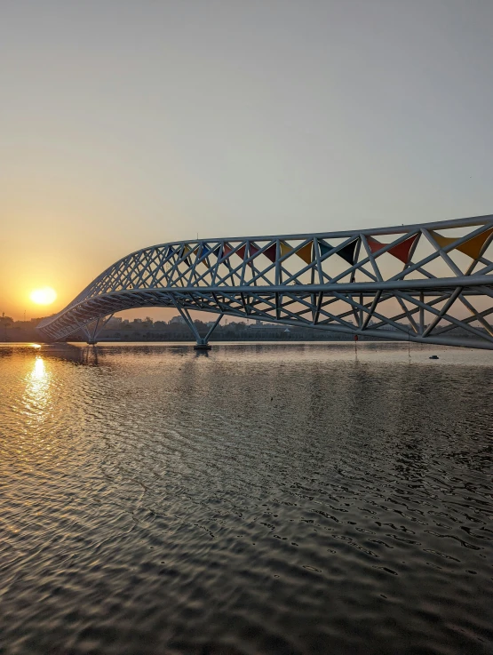 a metal bridge going across the water with a sun behind it