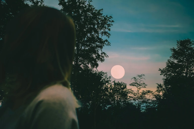 a woman stands against a pink moon during a dusk