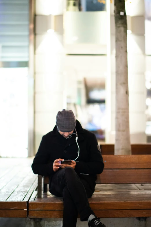 a young man in an overcoat sits on a bench and looks at his cellphone