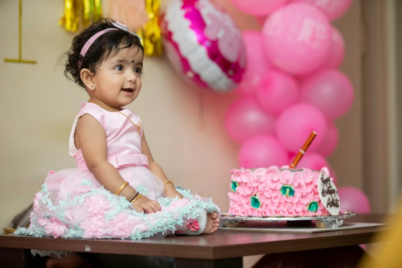 a baby girl wearing a pink dress sitting at a table in front of a birthday cake