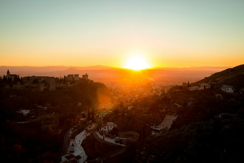 the sun rising over a city with trees and hills