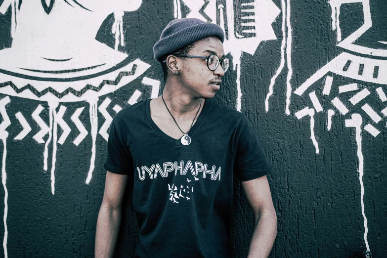 young man in black t - shirt standing by a wall covered in graffiti