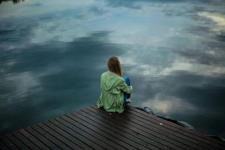 girl in green sitting on pier by water and cloudy sky