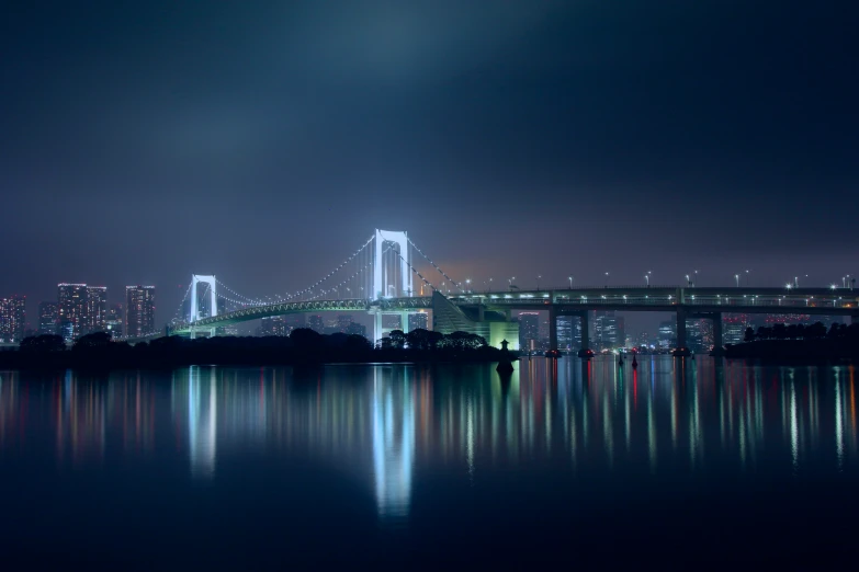 a city lights up over the water and under a bridge at night