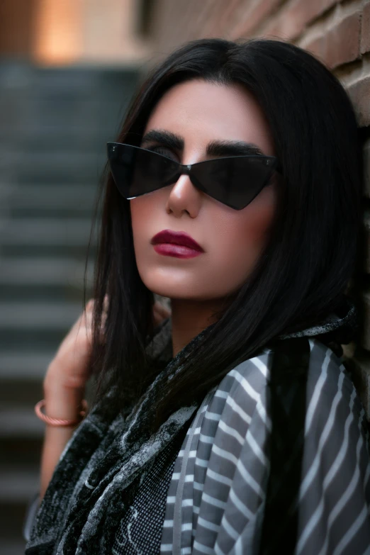 a woman with dark hair wearing sunglasses poses by a wall