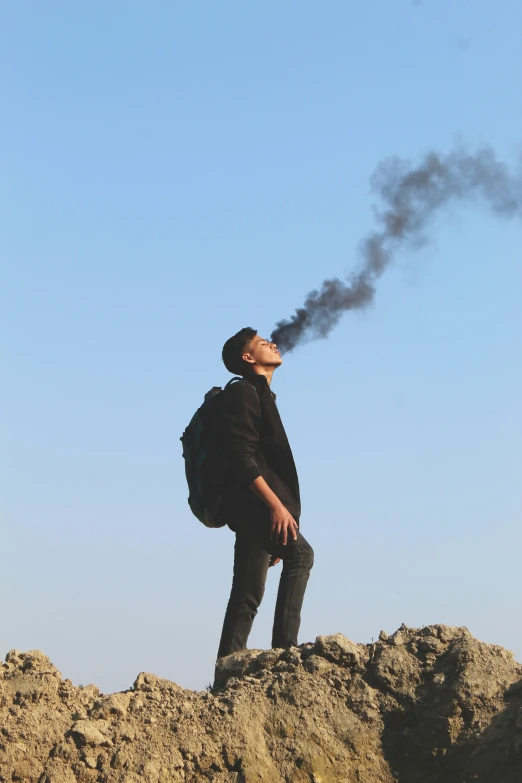 a man on a rocky area holding a backpack looking up at smoke coming from his mouth