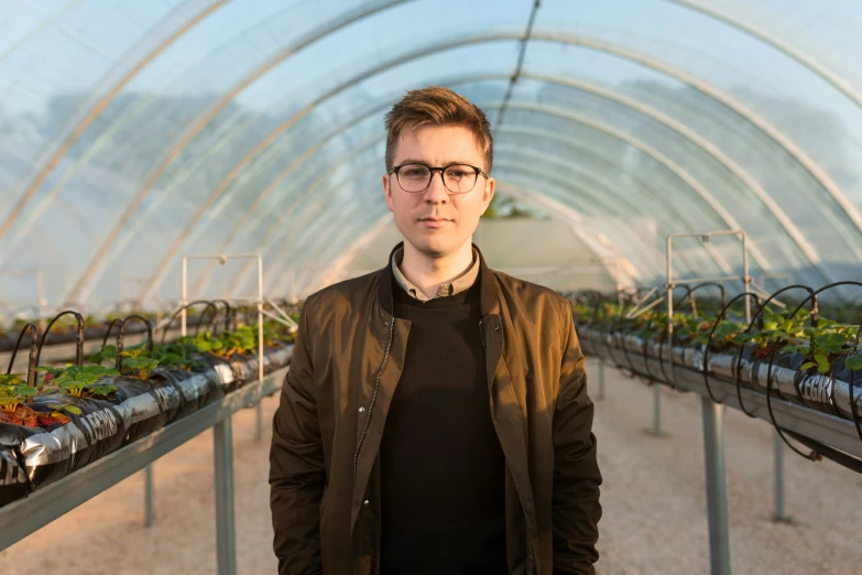 a man in glasses and a leather jacket in a greenhouse with many plants